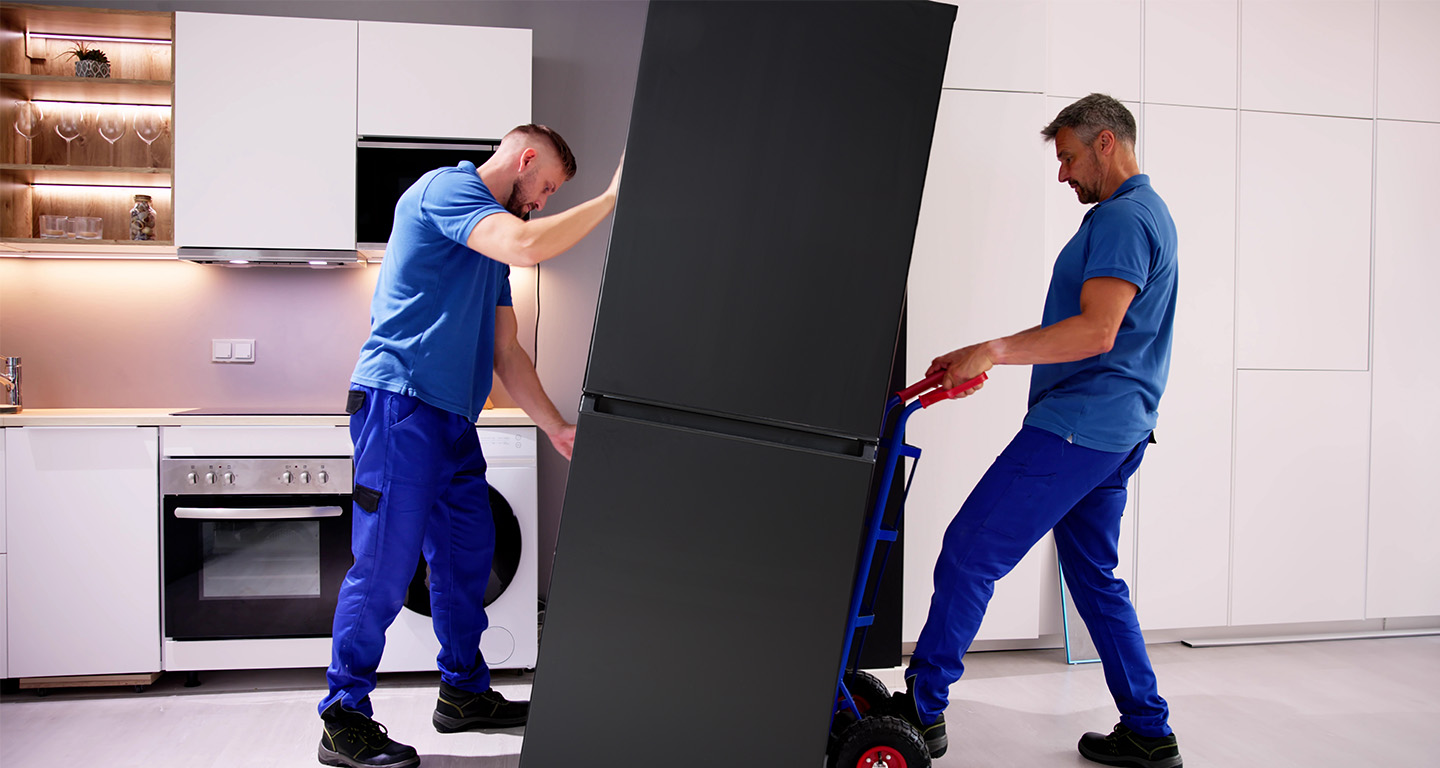 Two men with a two-wheeled dolly removing a refrigerator from a kitchen.