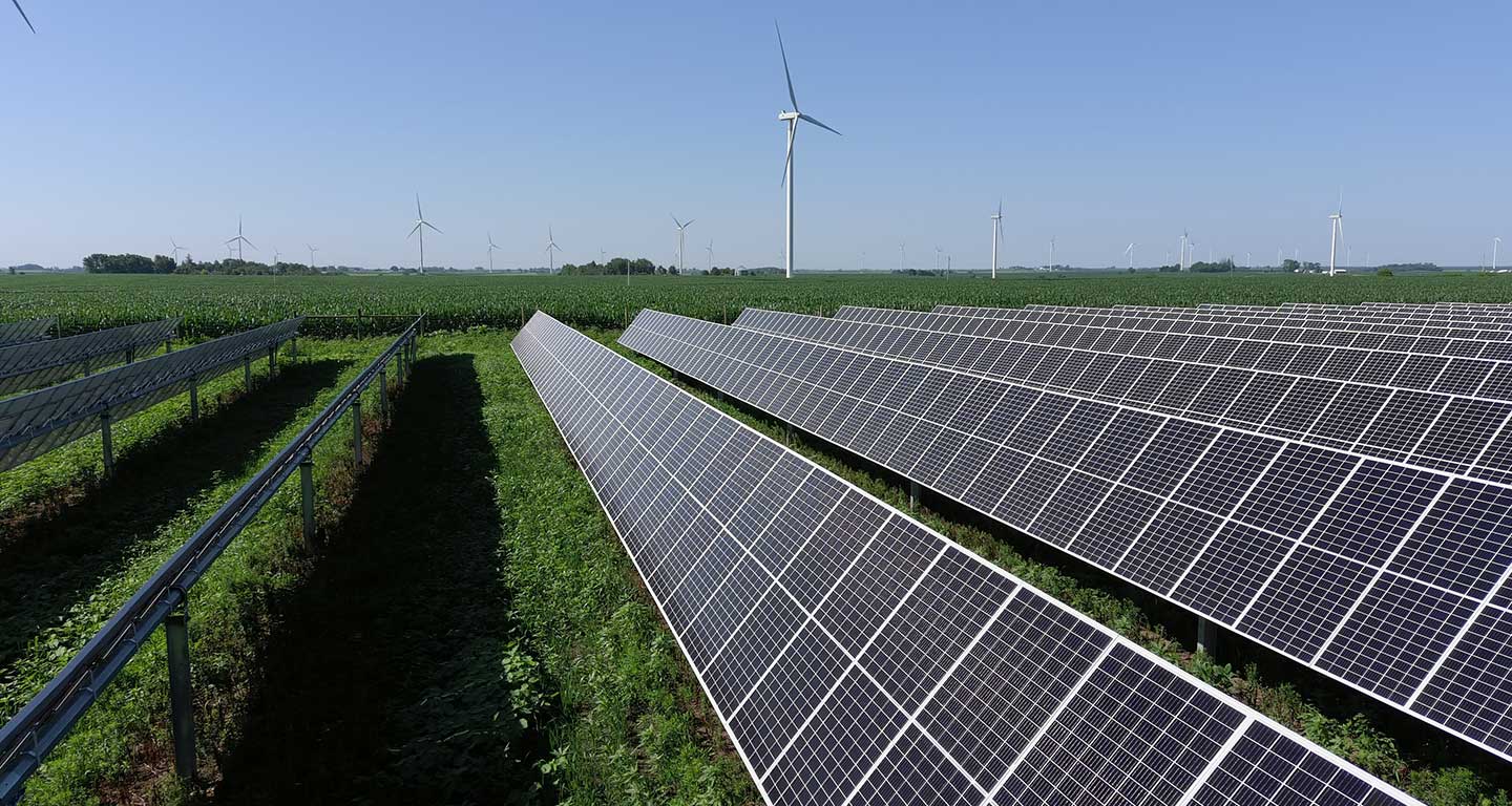 Side view of rows of solar panels, wind turbines in background