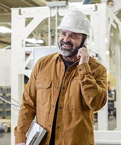 A man wearing a tan, long-sleeved shirt and a hardhat carrying a clipboard and speaking into a mobile phone, standing in an industrial warehouse setting.