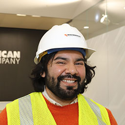 Smiling bearded worker with dark chin length hair wearing a safety vest and hard hat