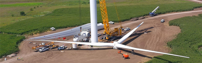 A wind turbine tower work site with several trucks and crews. The blades and nacelle have been removed for repowering. New blades awaiting installation are in the background.