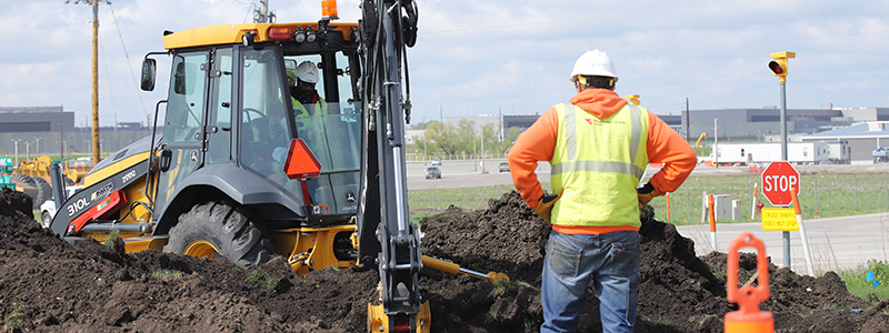 An excavator digging through a mound of black soil alongside a roadway while a MidAmerican employee wearing a high-visibility vest and hardhat supervises from a safe distance.
