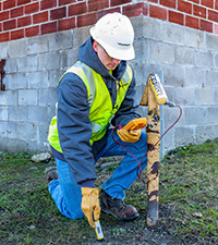 A MidAmerican employee wearing a high-visibility vest and hardhat kneels as he performs a gas leak test on an exterior gas pipe