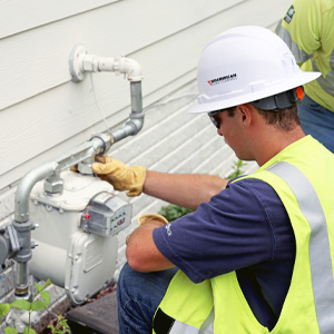 A MidAmerican employee wearing gloves, hardhat, high visibility vest and safety glasses installs a gas meter  outside of a residence.