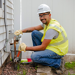 A smiling MidAmerican employee wearing a high-visibility vest, hardhat, safety glasses and gloves kneeling and using a crescent wrench on a pipe connecting to a residential gas meter