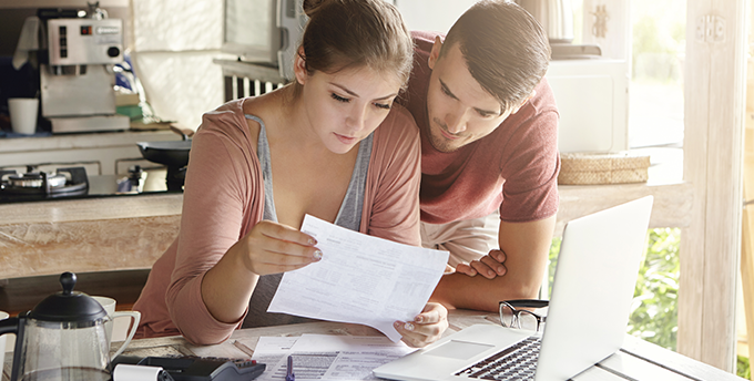 A couple looking over a bill, a laptop and some paperwork at a kitchen table