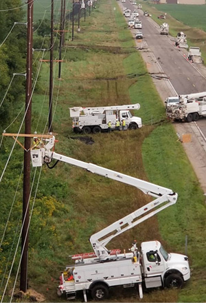 A vertical photo of several MidAmerican vehicles and crews replacing transmission poles along a roadside ravine in Illinois. A bucket truck with boom extended is in the foreground, additional trucks behind it and lining th.e roadway.