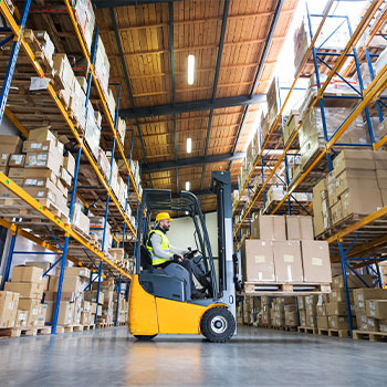 Man driving a forklift on the floor of a warehouse with a pallet of boxed goods lifted off the ground 