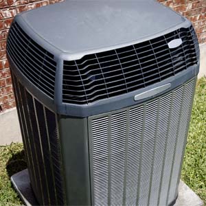 A grey air conditioner unit on a mounting pad viewed in three-quarters overhead and front with edges of lawn visible around the mounting pad and the exterior wall of a house behind