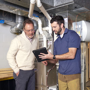 HVAC technician showing a customer a digital tablet while standing in front of a furnace in a residential basement
