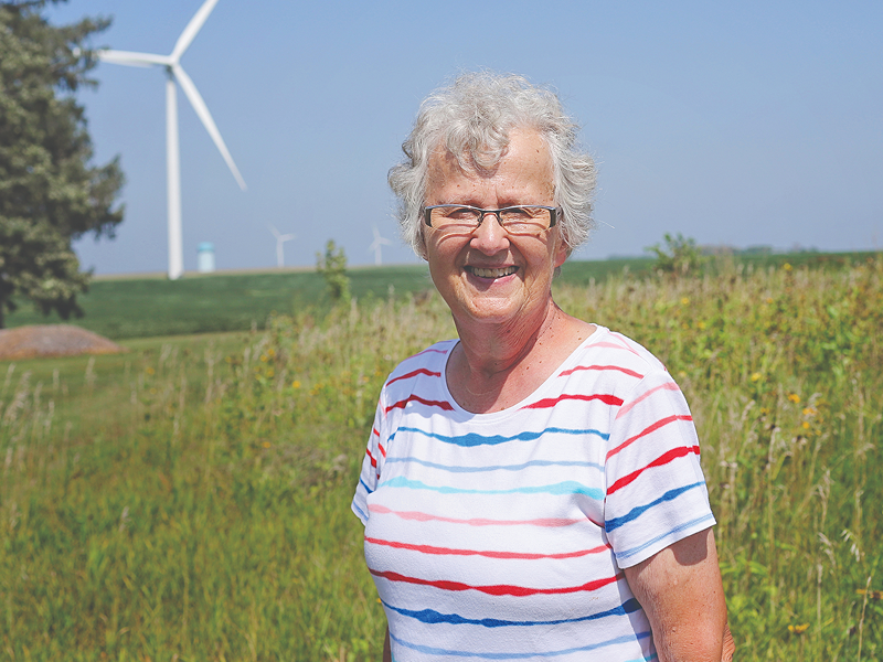 Evie, an older woman with white hair and glasses, wearing a multicolored striped shirt smiling with farmland and wind turbines in the background