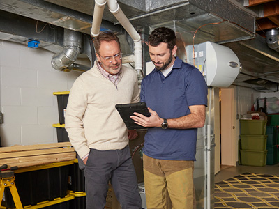 Two men in the basement of a home, looking at a tablet with a furnace in the background