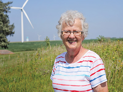 Evie, an older woman with white hair and glasses, wearing a multicolored striped shirt smiling with farmland and wind turbines in the background