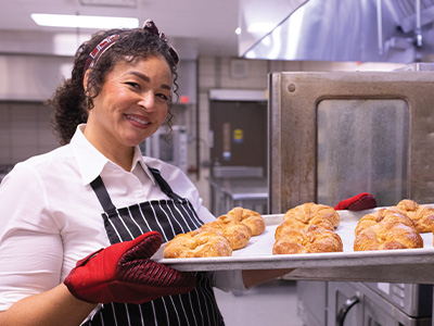 A woman with dark hair, a white shirt, and striped apron smiles while she removes a tray of freshly-baked croissants from a commercial oven