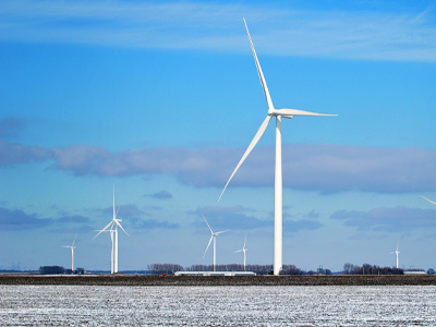 Horizontal image of a MidAmerican wind farm with six white wind turbines in a snowy crop field, visible against a partly cloudy blue sky and farm buildings in the background.