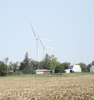 A harvested field, farmhouse and barn in the foreground with two wind turbines visible behind them