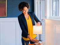 A casually-dressed woman in a living room, with her hand inside of the shade of a small lamp on a table, adjusting a bulb that has just been lit