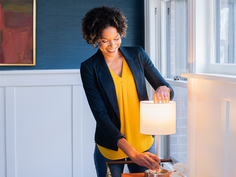 A casually-dressed woman in a living room, with her hand inside of the shade of a small lamp on a table, adjusting a bulb that has just been lit