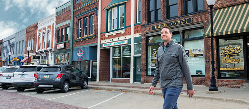 A man in a grey quarter zip crosses the street in front of a strip of small businesses