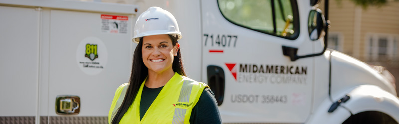 A smiling MidAmerican employee wearing a hardhat and visibility safety vest standing in front of a MidAmerican truck.