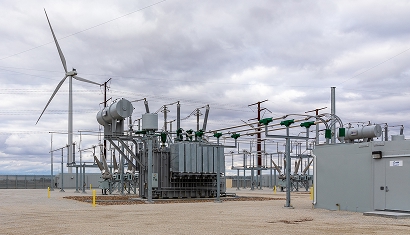 An electric substation with transmissoin wires an a wind turbine in the background against a bright, cloudy sky.