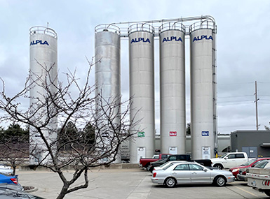Five silos marked ALPLA behind a parking lot against an overcast sky