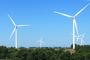 A  photo of white wind turbines standing on a green field lined with trees, with other turbines from the same windfarm visible in the background against a bright blue sky.
