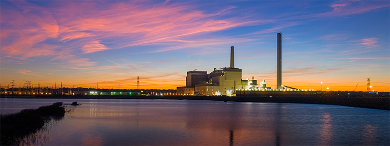 Wide format photo taken across the river of the Walter Scott, Jr. Energy Center with the sun setting behind it and colors reflecting in the water.
